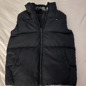 Svart dunväst från Tommy Hilfiger - Svart dunväst från Tommy Hilfiger med hög krage och diskret logga på bröstet. Västen har två sidofickor och dragkedja framtill. Insidan har broderad Tommy Hilfiger-text vid nacken. Perfekt för lager-på-lager under kyliga dagar.