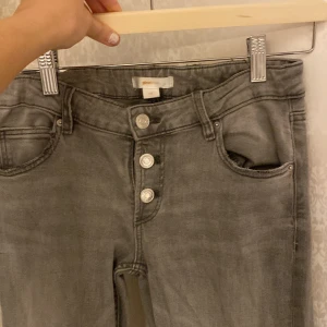 Gråa jeansbyxor från Gina Tricot - Snygga grå jeansbyxor från Gina Tricot med låg midja och utsvängda ben. Byxorna har fyra synliga silverknappar framtill, klassiska femficksdetaljer. Perfekta för en chill och trendig look. Passar kanske nån mellan 158-165