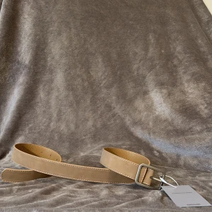 Beige bälte - Stilrent beiget bälte med matt silverfärgat spänne. Bältet har en klassisk, rak form och är gjort i skinn med ljusbrun insida. Perfekt accessoar för att lyfta din outfit. 100cm
