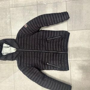 Grå/svart dunjacka med huva 154 cm - Svart/grå dunjacka från colmar med huva och två dragkedjeförsedda fickor framtill. Jackan har quiltad design och lång ärm, samt en colmar logga på armen. Perfekt för kyliga dagar och har en stilren look.