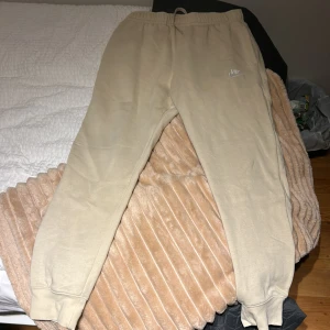 Beige mjukisbyxor från Nike - Beiga mjukisbyxor från Nike med broderad logga på vänster lår. Byxorna har resår och snörning i midjan samt muddar vid bensluten. Tillverkade i mjukt bomullsmaterial för extra komfort. Perfekta för chill eller träning.