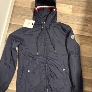 Mörkblå vindjacka från Moncler - Snygg mörkblå vindjacka från Moncler med huva och dragkedja framtill. Jackan har vita och röda detaljer på huvan, snörning och två fickor med tryckknappar. Klassisk Moncler-logga på ärmen och lätt, sportig känsla.