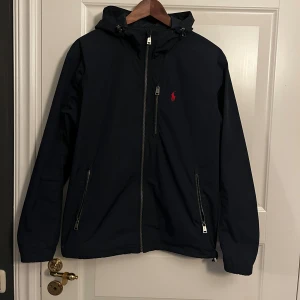 Ralph Lauren vindjacka - Marinblå vindjacka från Polo Ralph Lauren med huva, dragkedja framtill och två fickor med dragkedja. Jackan har röd broderad logga på bröstet och justerbar nederkant.