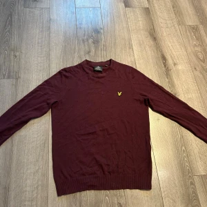 Vinröd tröja från Lyle & Scott - Vinröd långärmad tröja från Lyle & Scott med rund hals och gul broderad logga på bröstet. Tröjan är stickad och har ribbade muddar vid ärmslut och nederkant. Perfekt för lager-på-lager under hösten och vintern.