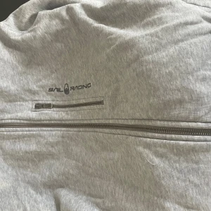 Grå jacka från Sail Racing - Snygg grå jacka från Sail Racing med två dragkedjefickor framtill och stilren logga på bröstet. Jackan har en sportig look och är perfekt för dig som gillar en clean och enkel stil.