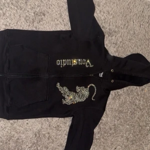 Svart Von Dutch hoodie med paljetttiger - Svart hoodie från Von Dutch med dragkedja, huva och fickor framtill. På bröstet finns en glittrig tiger i paljetter och Von Dutch-loggan i silver. Perfekt statement-plagg för dig som gillar unika detaljer och streetstyle.