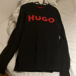 Svart tröja med röd HUGO-logga - Svart långärmad tröja från Hugo Boss med stor röd HUGO-logga framtill. Klassisk passform med ribbade muddar vid ärmslut och hals. Perfekt för dig som gillar stilrena och trendiga plagg med tydlig logga.