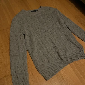 Grå kabelstickad tröja Polo Ralph Lauren - Säljer en klassisk grå kabelstickad tröja från Polo Ralph Lauren. Tröjan har rund halsringning, ribbade muddar och är långärmad. Perfekt för lager-på-lager och snygg till jeans eller kjol.
