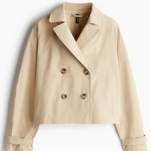 Beige trenchcoat från H&M - Säljer en beige croppad trenchcoat från H&M med dubbelknäppning och klassisk krage. Jackan har långa ärmar med knappar vid ärmslut och snygg detalj i ryggen. Perfekt för dig som vill ha en stilren och trendig look. Använd några gånger men ser ut som nu då den inte har några difekter. Skriv om ni vill ha fler bilder. Pris kan diskuteras.