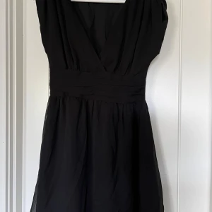 Svart klänning  - Svart kortklänning med v-ringad hals. Klänningen har markerad midja och är ärmlös, med lätt rynkade detaljer upptill. Notera att sömmen uppe på ena axeln gått upp, går att sy för den som kan. 
