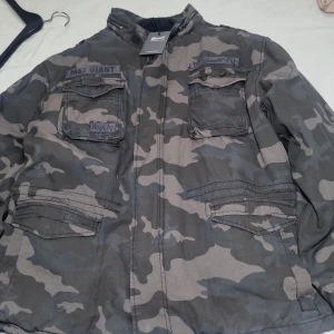 Camouflage parkajacka från Brandit - Säljer en grå och svart camouflage parkajacka från Brandit, modell M65 Giant. Jackan har flera stora fickor framtill, dragkedja och knappar, samt ett varmt grått fleecefoder på insidan. Perfekt för kalla dagar och riktigt snygg streetstil.