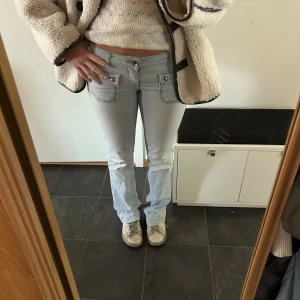Ljusblå bootcut jeans med fickdetaljer - Säljer ett par ljusblå jeans med låg midja och bootcut-ben. Jeansen har stora fickor framtill med lock och knappar, samt klassiska bakfickor. Snygga kontrastsömmar och coola detaljer som ger en trendig look. Perfekta för dig som gillar 00-talsvibbar.