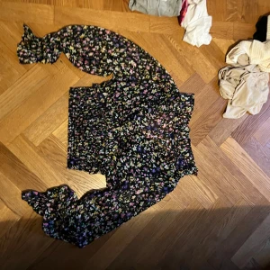 Svart blommig blus med puffärm - Säljer en svart blus med färgglatt blommigt mönster i lila, rosa, grönt och gult. Blusen har långa puffärmar med resår vid ärmslut och smockad midja för en snygg passform. Materialet är lätt och luftigt, perfekt för dig som gillar romantisk stil.