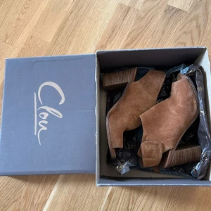 Bruna mocka stövletter från Clou - Snygga bruna stövletter i mocka från Clou med öppen tå och bred klack. Skorna har en stilren design med kardborreknäppning vid hälen och en plattformsinspirerad sula. Perfekta för dig som vill ha en trendig och bekväm sko med lite höjd.