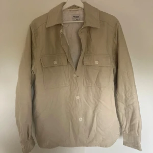 Beige overshirt från Weekday - Stilren beige overshirt från Weekday med quiltat foder. Skjortan har två stora bröstfickor med knappar, klassisk krage och långa ärmar. Perfekt lager-på-lager-plagg med snygg, enkel design och bekvämt material. Storlek S men passar dig som är runt 180cm. Knappt använd och i bra skick!