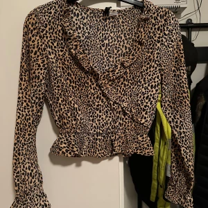 Leopardmönstrad blus från DIVIDED strl: 32 - Säljer en beige och svart leopardmönstrad blus från DIVIDED i storlek 32. Blusen har omlott-look med volangdetaljer vid halsringning, midja och ärmslut. Långärmad och croppad passform, tillverkad i mjuk polyester. Perfekt för dig som gillar djurmönster och volanger!