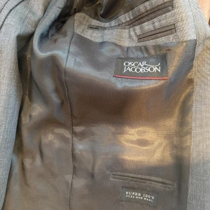 Grå kostym från Oscar Jacobson i ull - Snygg grå kostym från Oscar Jacobson i Super 100's ren ull. Kavajen har klassisk passform med tre knappar vid ärmslut och diskret mönster. Byxorna är raka och matchar kavajen perfekt. Perfekt för dig som vill ha en stilren och tidlös look.