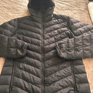 Svart pufferjacka från Haglöfs M - Svart pufferjacka från Haglöfs i storlek M. Jackan har snygga diagonala quiltade paneler, hög krage och två dragkedjeförsedda fickor. Materialet är syntet och jackan har en stilren look med diskret logga på bröstet. Perfekt för kalla dagar.