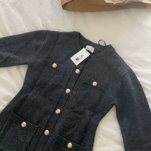 Mörkgrå figursydd kofta med guldknappar från hm - Mörkgrå figursydd kofta från H&M med snygga guldiga knappar framtill och på fickorna. Modellen är rak med rund halsringning, långa ärmar och två fickor framtill. Perfekt för dig som gillar klassisk men trendig stil.