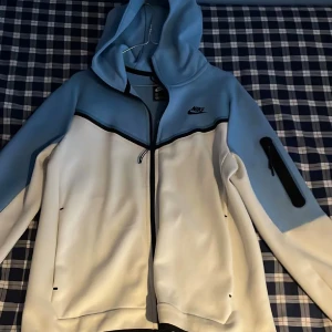 Nike tech blåvit vindjacka med huva - Nike vindjacka i ljusblått och vitt med svart detaljlinje över bröstet. Jackan har huva, dragkedja framtill och en extra ficka med dragkedja på ärmen. Perfekt för dig som gillar sportig och clean stil.