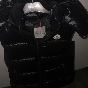 Svart dunväst från Moncler - Svart glansig dunväst från Moncler med hög krage och dragkedja framtill. Västen har två fickor med dragkedja och klassisk Moncler-logga på bröstet. Perfekt för lager-på-lager och riktigt snygg till streetwear. Använd Bara 2 Gånger Pga Den Inte Passar. Passar S och M