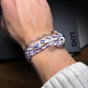 Flätat armband med ankare - Säljer detta unika armband från Blvckout med marin känsla, gjort av vitt, blått och rött flätat rep och en silverfärgad ankare som lås. Snygg detalj med märkets logga ingraverad på ankaret. Perfekt för en som gillar stilrena smycken. Nypris ca 400kr, kom med prisförslag⚓️🌊💍.
