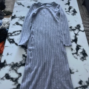 Grå ribbad långklänning - Snygg grå långklänning med ribbat mönster och långa ärmar. Klänningen har en enkel, rak passform och är tillverkad i ett mjukt stickat material som känns skönt mot huden. Perfekt för dig som gillar stilrena och bekväma plagg med lite edge.