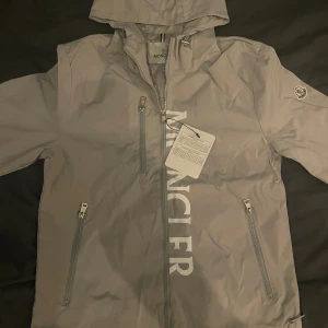 Grå vindjacka - Snygg grå vindjacka med huva och dragkedja framtill. Jackan har tre fickor med dragkedja, logga på ärmen och stort tryck på bröstet. Perfekt för dig som vill ha en stilren och sportig look