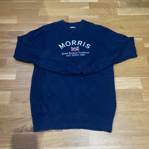 Mörkblå stickad tröja från Morris - Mörkblå stickad tröja från Morris med broderad logga och brittisk flagga på bröstet. Klassisk rund halsringning och ribbade muddar. Perfekt för dig som gillar stilren och tidlös design.