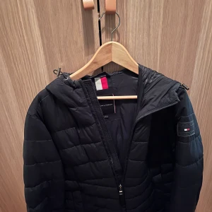 Svart pufferjacka från Tommy Hilfiger - Svart pufferjacka från Tommy Hilfiger med huva och dragkedja. Jackan har quiltad design och klassisk logga på ärmen. Perfekt för kyliga dagar och har en stilren look som funkar till det mesta.