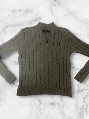  tröja från Polo Ralph Lauren - Kabelstickad grå tröja från Polo Ralph Lauren med halv dragkedja och broderad logga på bröstet. Tröjan har ribbade muddar och hög krage, perfekt för lager-på-lager. Klassisk och stilren design i mjukt material.