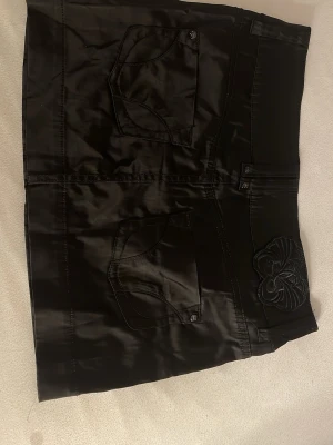 Vintage Gina tricot kjol - Svart kortkjol från Gina Jeans med två framfickor och dekorativ broderad blomdetalj vid midjan. Kjolen har en glansig finish och markerade sömmar som ger en cool look. Perfekt att styla med en enkel topp eller hoodie.