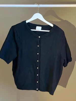 Svart kortärmad kofta med pärlknappar - Stilren svart kofta med korta ärmar och pärlknappar framtill. Koftan har rund halsringning och är stickad i ett mjukt material. Perfekt att slänga över en topp för en enkel men snygg look. Köpt från H&M, helt oanvänd🩶