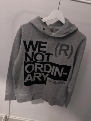 Grå hoodie från Jack & Jones - Snygg grå hoodie från Jack & Jones med stor svart text 'WE (R) NOT ORDINARY' på framsidan. Klassisk huva och ribbade muddar vid ärmar och nederkant. Perfekt för en avslappnad och trendig stil.
