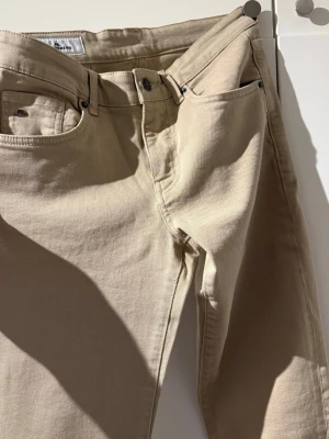 Beige jeans från J.Lindeberg 30/30 - Snygga beige jeans från J.Lindeberg i klassisk femficksmodell. Jeansen har normal passform och är tillverkade i mjuk bomull med en stilren look. Perfekta för dig som vill ha ett par ljusa jeans med enkel design.