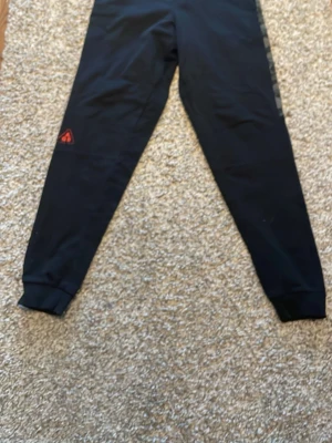 Svarta mjukisbyxor AG9 - Svarta mjukisbyxor från AG9 med smal passform och ribbade muddar vid bensluten. Byxorna har en diskret logga i rött på ena benet och är tillverkade i mjukt material som känns skönt mot huden. Perfekta för chill eller träning.