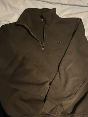 Svart Nike half zip sweatshirt - Svart sweatshirt från Nike med halv dragkedja och hög krage. Tröjan har diskret Nike-logga broderad på bröstet och ribbade muddar vid ärmslut och nederkant. Perfekt för en sportig och avslappnad stil.