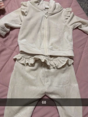 Krämvitt velourset med volanger - Mysigt loungewear-set i mjuk krämvit velour med volangdetaljer på axlar och midja. Setet består av en långärmad ziptröja med huva och matchande byxor med volangkant. Perfekt för chill dagar hemma.