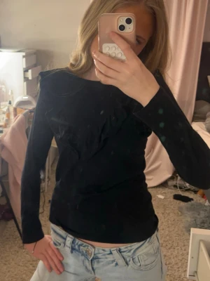 Svart långärmad topp med volang - Snygg svart långärmad topp med volangdetalj framtill. Toppen har en rund halsringning och sitter figurnära. Perfekt att styla med jeans eller kjol för en trendig look.