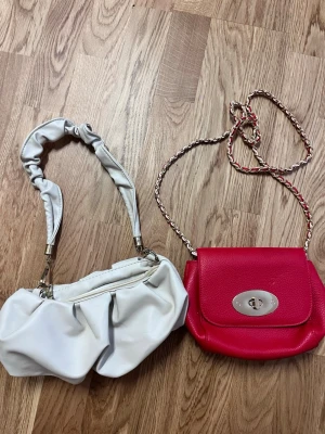 Två trendiga handväskor i vitt och rött - Säljer två snygga handväskor. Den ena är beig med scrunchie-handtag och mjuk form, den andra är röd med kedjerem . Båda för 80 kr