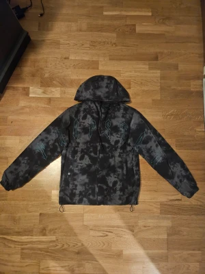 Svart och grå vindjacka från Trapstar - Säljer en svart och grå mönstrad vindjacka från Trapstar med huva och dragkedja. Jackan har ett unikt batikliknande mönster och blå textdetaljer över bröstet och ärmarna. Justerbar nederkant med snörning och långa ärmar. Perfekt för dig som vill sticka ut.