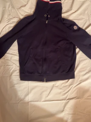 Mörkblå hoodie från Moncler - Snygg mörkblå hoodie från Moncler med dragkedja framtill och fickor på magen. Huvan har vita och röda detaljer upptill och är försedd med snörning. Moncler-logga på ärmen ger en exklusiv touch. Perfekt för dig som gillar stilrena och sportiga plagg.