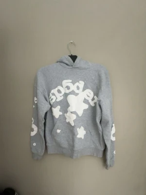 Sp5der hoodie  - Säljer en grå hoodie från Sp5der med stora vita tryck och stjärnor på både fram- och ärmar. Hoodien har en klassisk huva och ribbade muddar. Perfekt för dig som gillar streetwear och vill sticka ut med cool grafik.