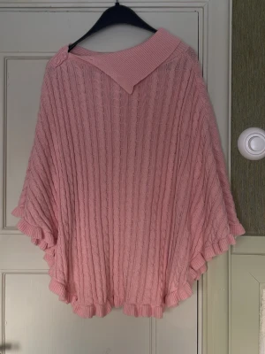 Rosa stickad poncho med knappar - Säljer en söt rosa stickad poncho med kabelstickat mönster och volangkant. Ponchon har en bred krage och två matchande knappar vid axeln för extra detalj. Perfekt för lager på lager och ger en mysig vibe till din outfit.