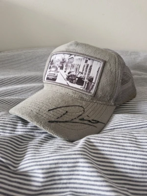 Emilio pendenza signerad av dino beganovic - Säljer en grå keps med mesh på sidorna och baksidan. Framtill finns ett svartvitt fototryck av en gata med bilar och hus, samt en svart signatur på skärmen. Kepsen har en böjd skärm och justerbar passform bak. Perfekt för dig som gillar streetstyle och unika detaljer. Kepsen är signerad av blivande f1 förare