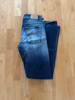 Blå jeans från Nudie Jeans, W29 L32 - Säljer ett par klassiska blå jeans från Nudie Jeans med snygg slitning och kontrastsömmar. Modellen har raka ben och fem fickor, samt Nudie-logga på bakfickan. Perfekta för dig som gillar en tidlös och avslappnad stil.