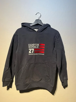 Hampton Republic hoddie - Mörkblå hoodie från i STORLEK 164Hampton Republic med tryck framtill i vitt och rött. Klassisk känguruficka och huva. Mjuk bomullskänsla och ribbade muddar vid ärmslut och nederkant. Perfekt för en avslappnad och stilren look.