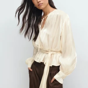 Krämvit omlottblus med knytband - Säljer en krämvit omlottblus med knytband i midjan och v-ringning. Blusen har långa, luftiga ärmar och volangdetalj nedtill. Perfekt för dig som gillar en romantisk och chic stil.