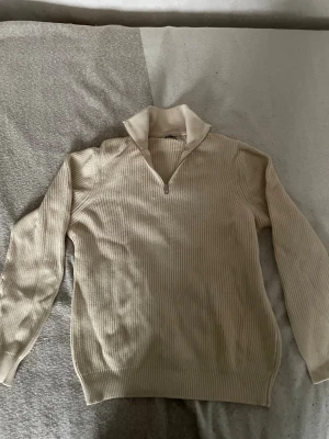 Beige ribbstickad tröja med dragkedja - Snygg beige ribbstickad half-zip med hög krage . Tröjan har långa ärmar och en relaxed fit, perfekt för coolingar. 