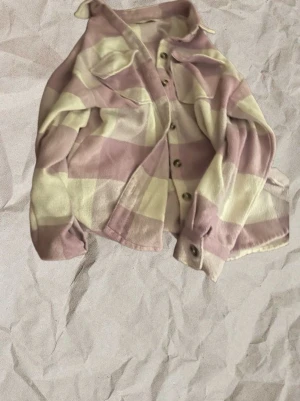 Rutig overshirt i lila och beige - Overshirt med breda rutor i lila och beige. Två bröstfickor med lock och knappar framtill. Skön och lite loose passform, perfekt att slänga över en t-shirt. Materialet känns mjukt och lite fleece-aktigt.